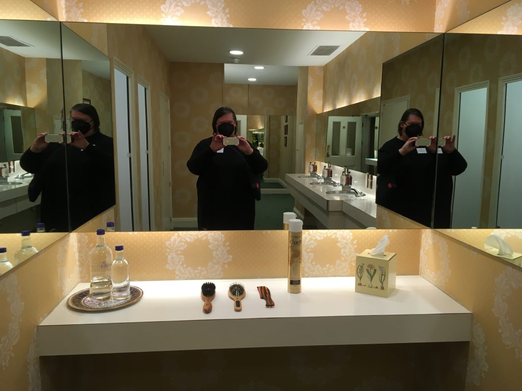 Mirror selfie in Buckingham Palace toilets, sinks in the background, general peachy-gold colour scheme, dyke wearing a black sack dress and Covid mask in the foreground. Brushes, a comb, water, tissues and hairspray are available for guests