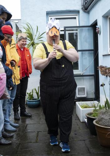 Fat dyke parades in a paper headdress of a swan's head, she plays the swanee whistle and is wearing dungarees. It is raining, people watch