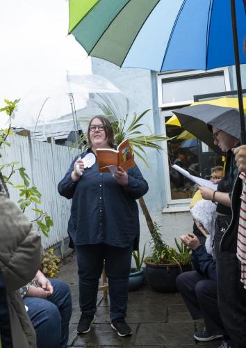 Fat dyke reading from her book Poundbury: A Queer Tour of Monarchy in the rain under an umbrella, people standing around