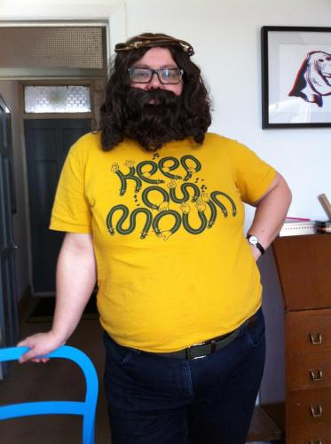 A dyke wearing a wig, beard and crown of thorns to look like Jesus or Allen Ginsberg, t-shirt, jeans, domestic setting