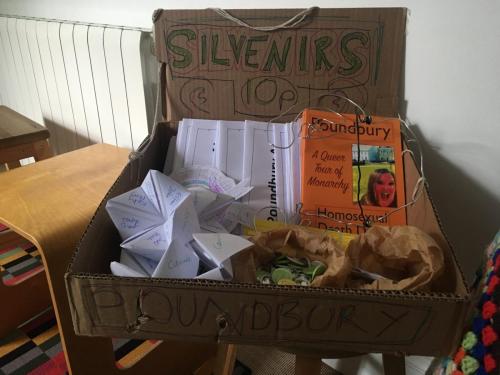 Cardboard tray featuring zines, badges, fortune tellers, bookmarks, stickers and copies of Poundbury: A Queer Tour of Monarchy by Homosexual Death Drive